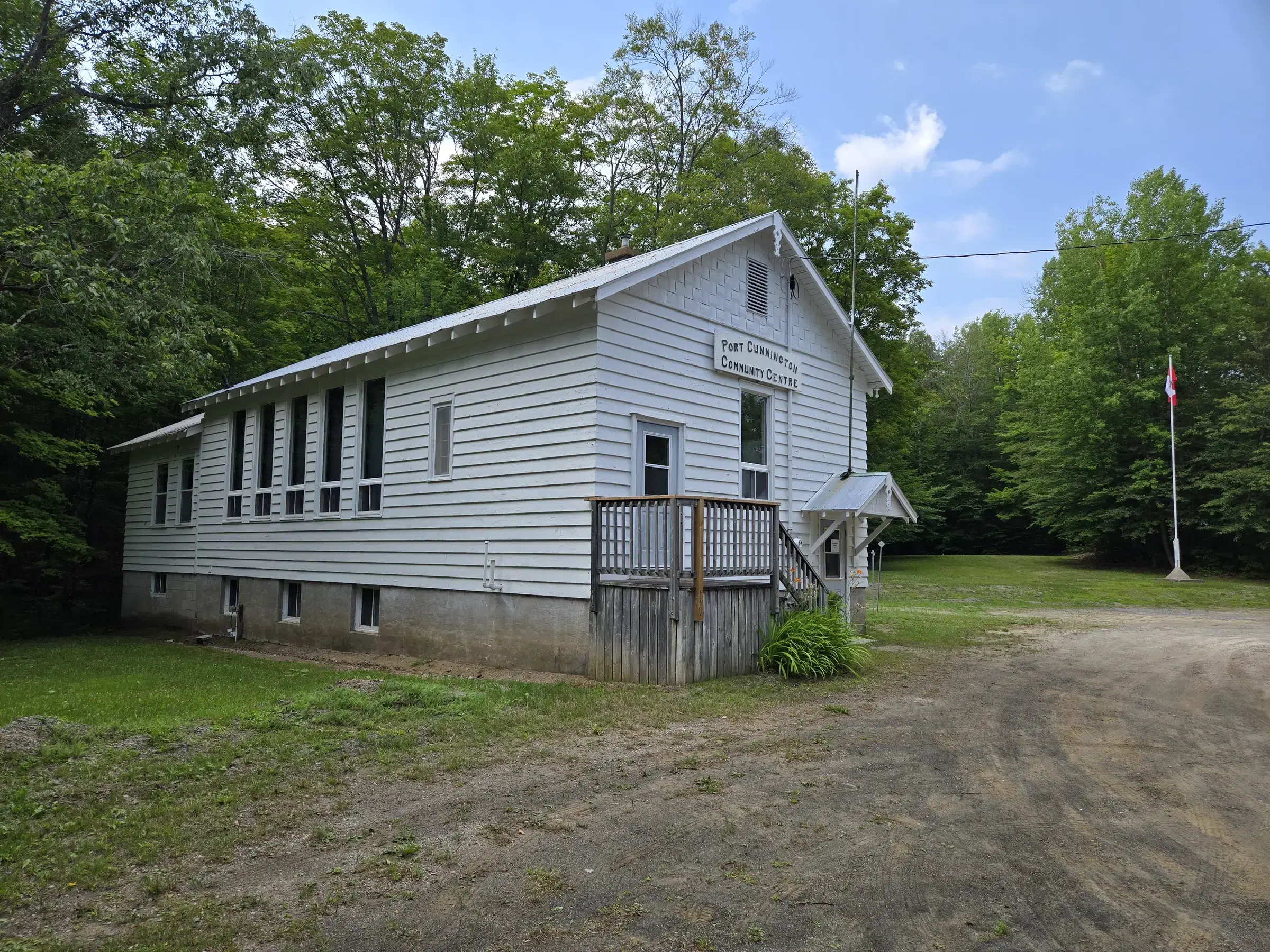 The community centre seen from the corner of the gravel lot, its long side wall in sun, with a Canadian flag on a pole to the right and mixed forest behind.