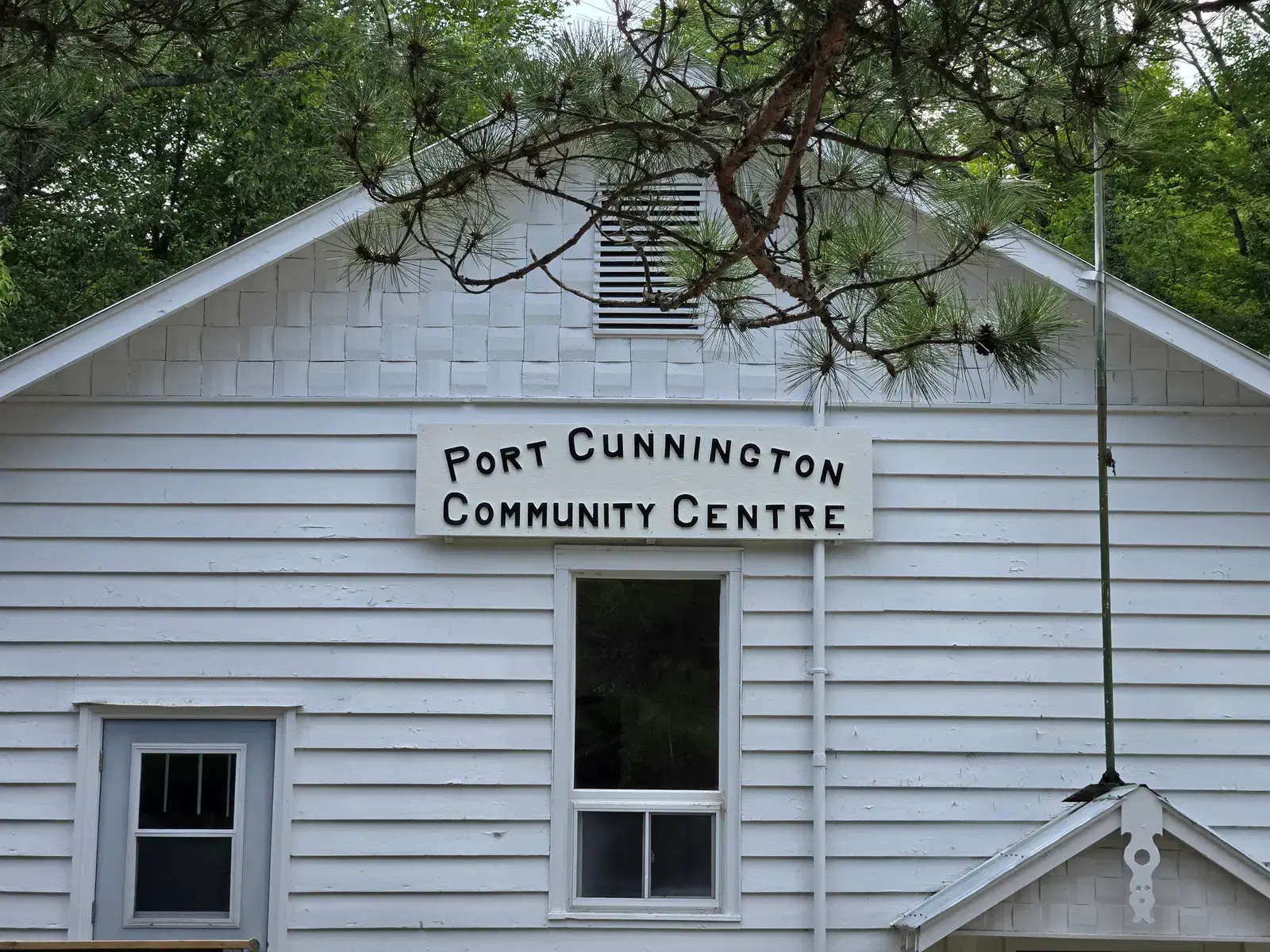 Close view of the hand-cut lettering on the sign reading 'Port Cunnington Community Centre', mounted on the clapboard below the gable.