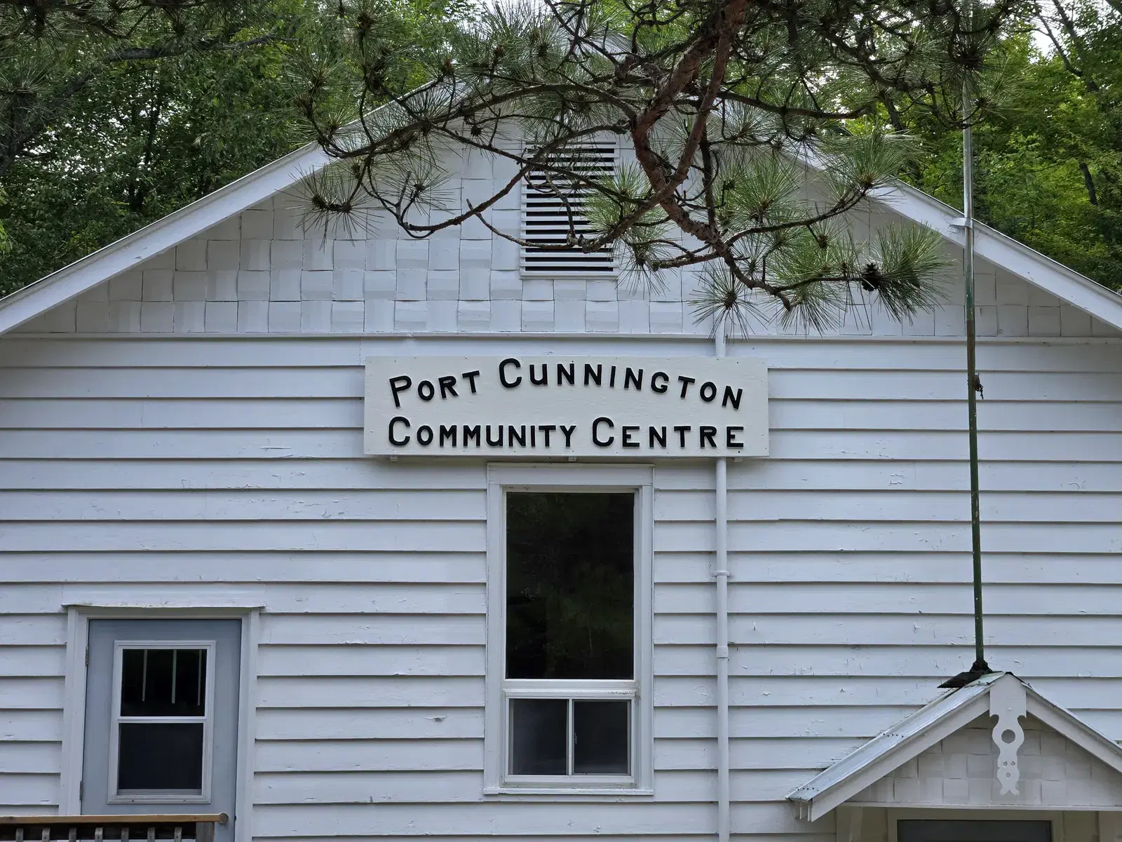 The gable end of the hall, its shingled peak and hand-lettered sign framed by a pine bough.