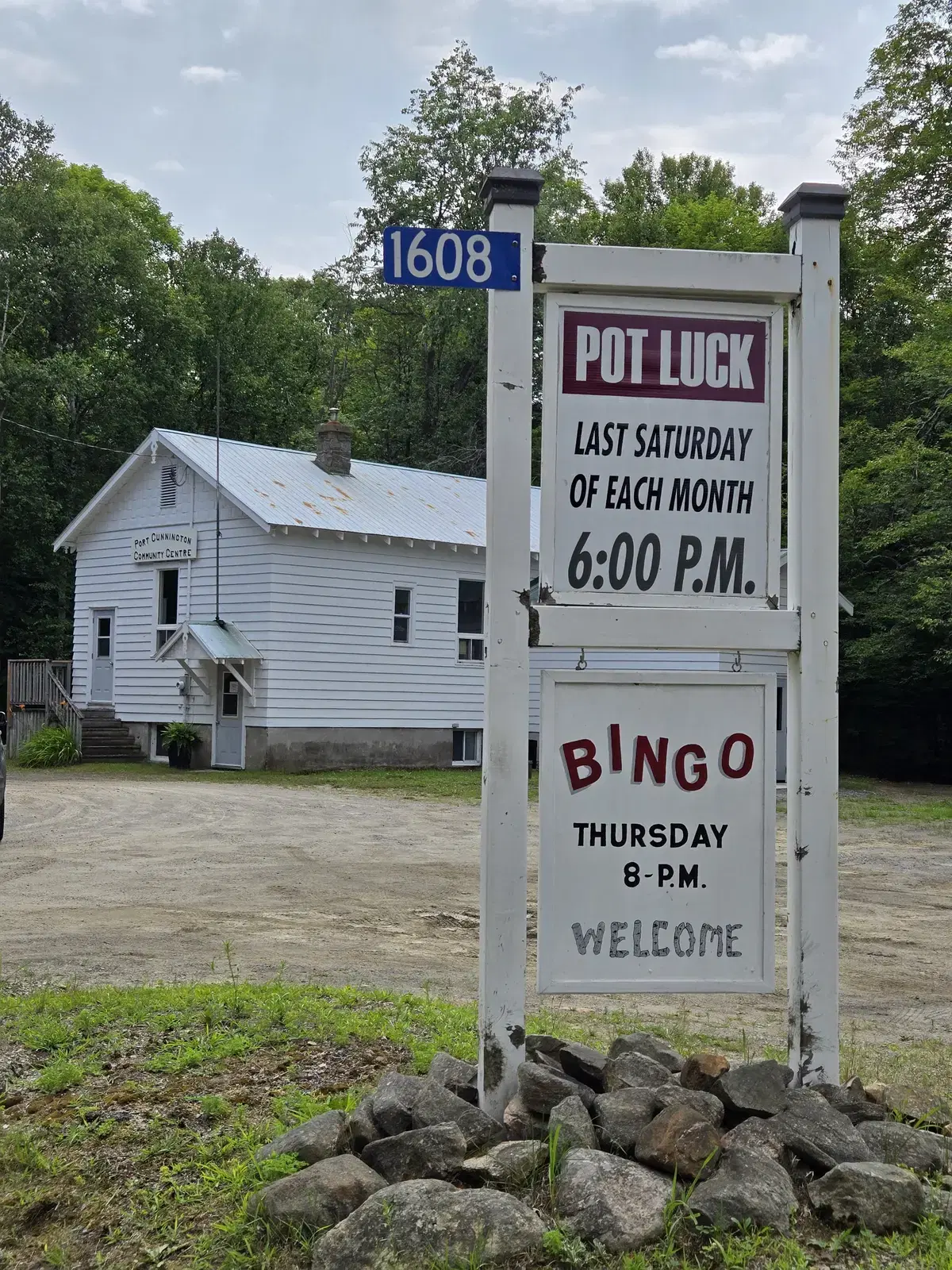 The roadside noticeboard at the end of the driveway, painted with 'Pot Luck, last Saturday of each month, 6:00 p.m.' above 'Bingo, Thursday 8 p.m. Welcome', with the hall behind it.